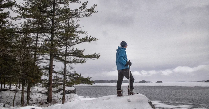 Person in a blue jacket standing on a snowy shore, looking out over a frozen lake with distant islands and surrounding pine trees.
