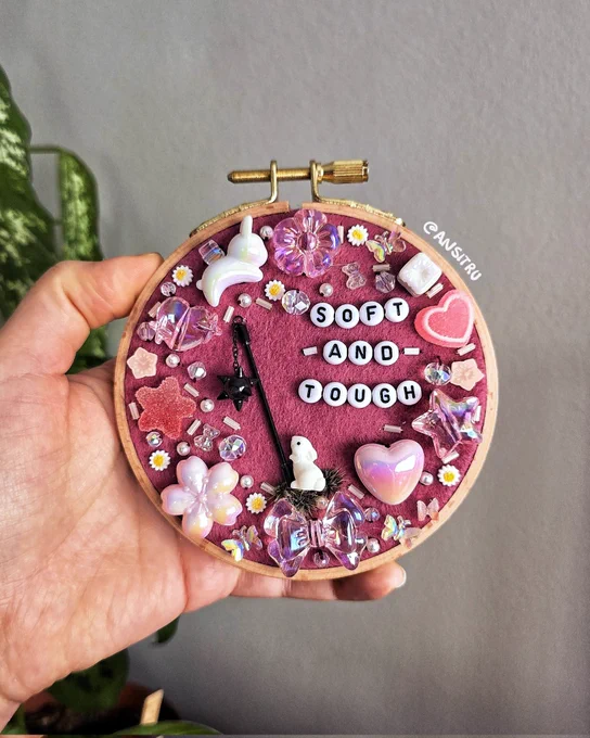 A hand holding a beaded embroidery hoop featuring a bunny holding a flail. The letter beads read "soft - and - tough"