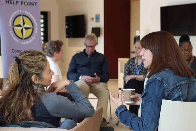Group of adults engaged in a discussion at a community help point, sitting in a circle with cups in hand.