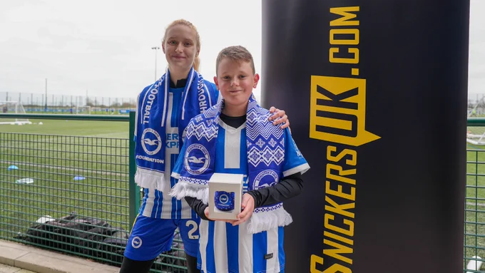 A boy and a girl in Brighton kits wear Albion scarfs and hold a Foundation bauble that was gifted to all the participants. They're stood next to a SnickersUK banner.