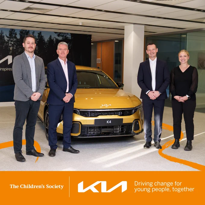 Four professionals stand around a yellow Kia car inside a modern showroom with The Children's Society branding visible.