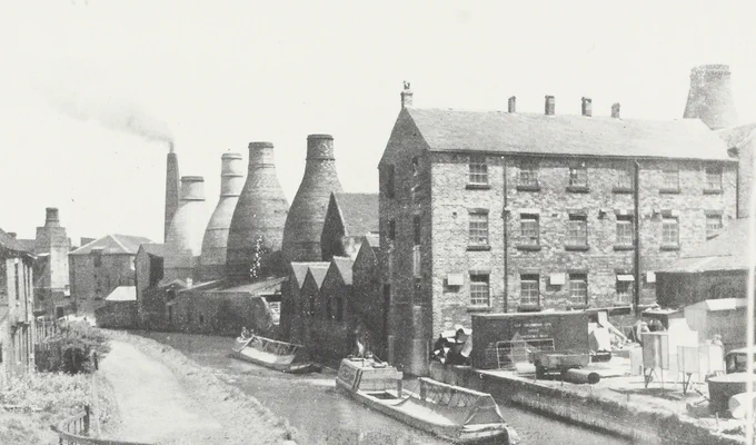 A historic black-and-white photograph of a canal-side pottery works, with bottle kilns, factory buildings, and narrowboats moored along the water.