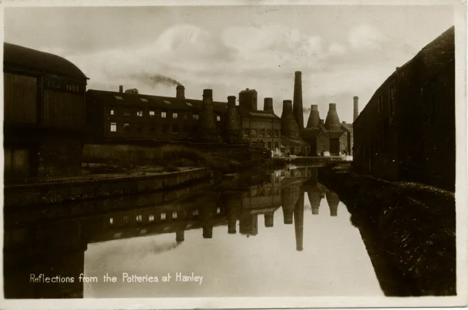 A historic photograph of the Potteries at Hanley reflected in a calm canal, showing bottle kilns, factory buildings, and chimneys lining the waterway.