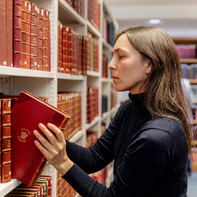 A woman with long brown hair pulls a red book from a bookshelf.