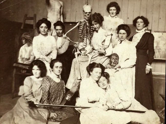 A vintage photograph depicts a group of women in Victorian-era attire, gathered in a studio setting. They are positioned around a standing human skeleton, with some women playfully interacting with it. The scene exudes a sense of curiosity and exploration, typical of a period art or science class. A mannequin is also visible in the background.