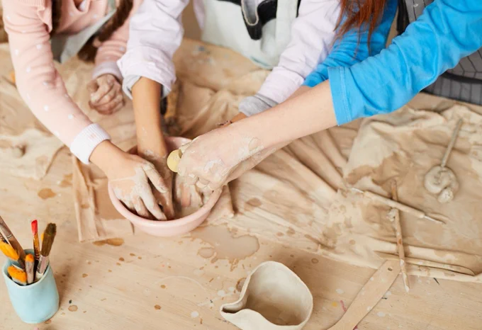 Hands shown covered in clay and being dipped into a bowl. Clay moulding tools, clay and clay pots surround them.