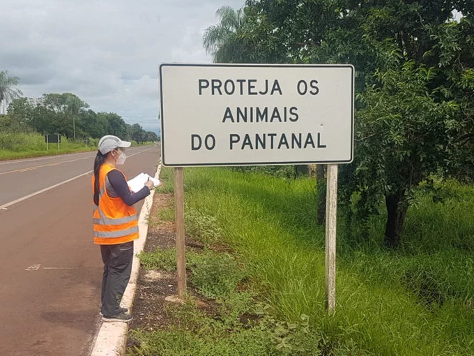 A woman with a clipboard next to a warning sign alongside a road. She is surveying the sign characteristics (size, wording etc)
