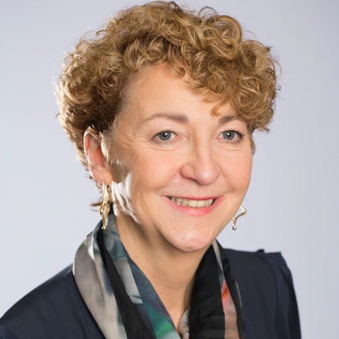 Woman with short curly blonde hair wearing a dark blazer and a multicoloured scarf against a plain background.