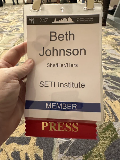 A woman’s hand holds an attendee badge for the AAS 247th winter conference. Text: Beth Johnson, she/her/hers, SETI Institute, Member. A red press ribbon hangs down from the bottom.