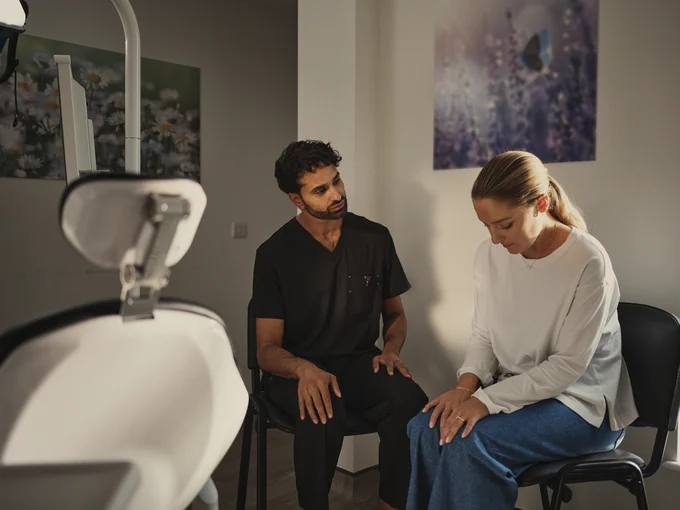 A healthcare professional in black scrubs consulting with a woman in casual clothes inside a softly lit dental practice.