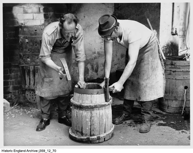A black and white photograph of two men standing on either side of a wooden barrel in a workshop. The man on the left is wearing an apron over a shirt and trousers, and the man on the right is wearing a hat and an apron. Both men hold the barrel steady with one hand and hold a hammer in their other hand. Barrels and tools are in the background.