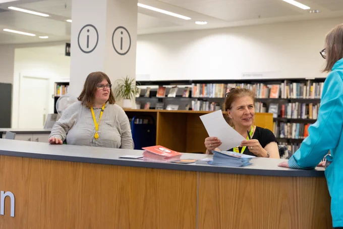 A librarian smiles as she greets a customer wearing a blue coat at an information desk.