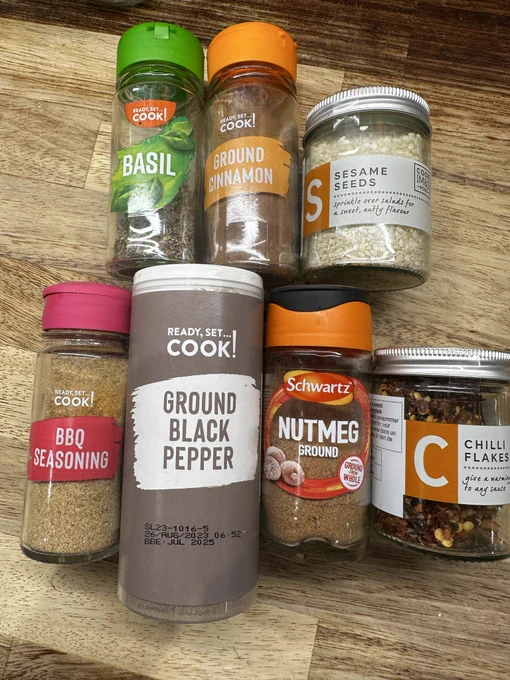 It’s a collection of 7 herbs and spices (and related) on a wooden bench/counter top. They are basil, ground cinnamon, sesame seeds, BBQ seasoning, ground black pepper, ground nutmeg and chilli flakes.
