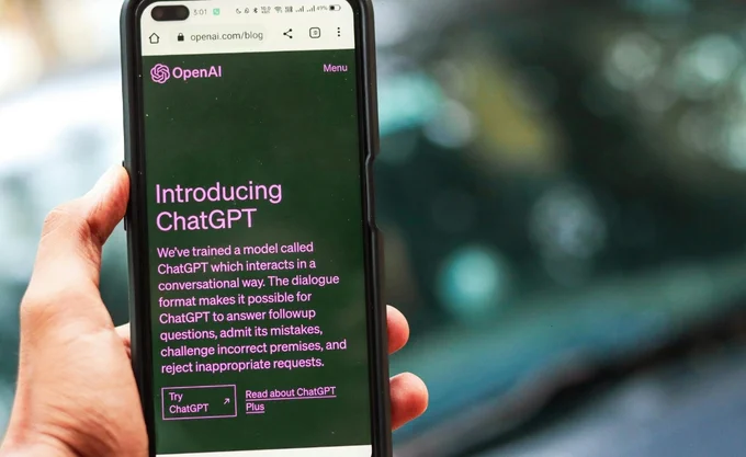A person holds a smartphone displaying a webpage from OpenAI. The screen features the title "Introducing ChatGPT" in purple text on a dark background, with a description of ChatGPT's capabilities. Two buttons at the bottom say "Try ChatGPT" and "Read about ChatGPT Plus." Blurry background with natural light.