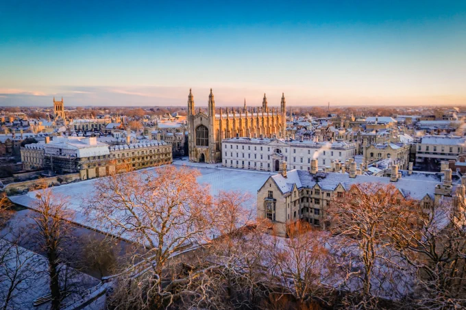 Aerial view of King's College Chapel in Cambridge, showing historic buildings covered in snow during a sunrise.