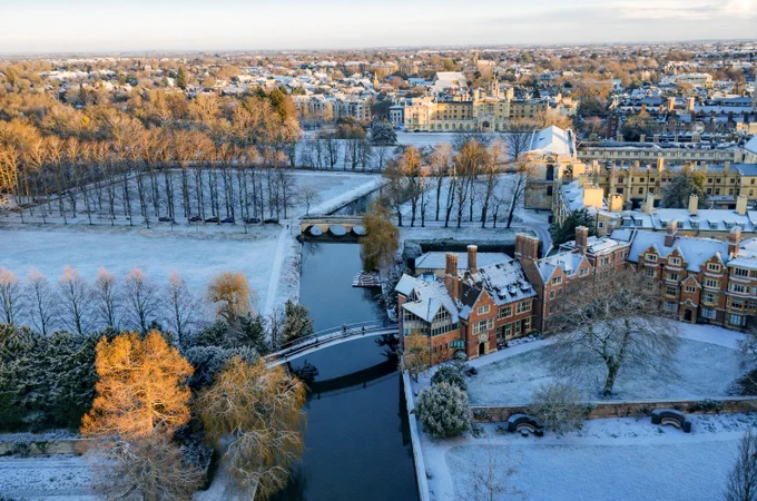Aerial view of Cambridge covered in snow, featuring the River Cam and several bridges connecting snow-covered landscapes and historic buildings.