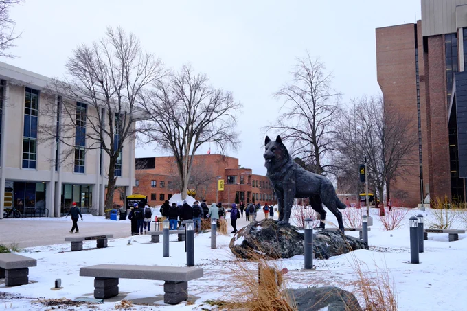 A snowy winter scene on campus featuring a large wolf statue in the foreground. Students in winter coats walk along a campus pathway between brick and modern academic buildings on the first day of classes.