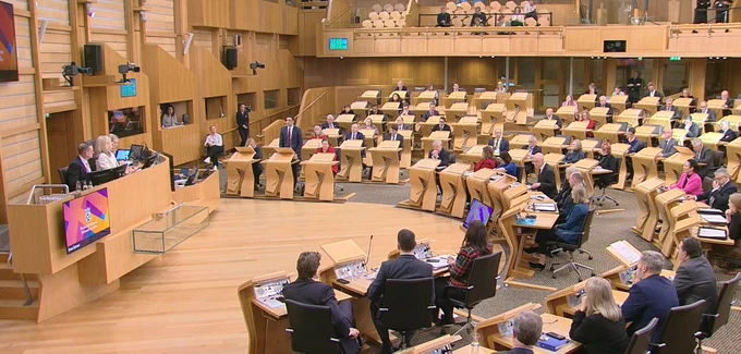 The Scottish Parliament Debating Chamber at First Minister's Questions. The chamber is full. Anas Sarwar MSP, Scottish Labour Party leader, is standing to ask a question, and the FM John Swinney MSP is getting to his feet to answer.