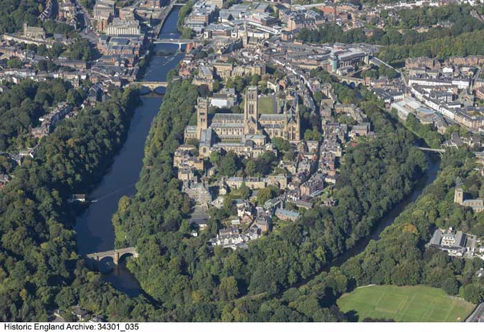 A colour photograph of an oblique aerial view of Durham Cathedral. The cathedral is in the centre of the photo. It is surrounded by green trees, with buildings along a road to the right and below it. The River Wear is seen clearly to the left, and wraps around the bottom and right of the city. At the top and edges of the photograph are buildings of the city.