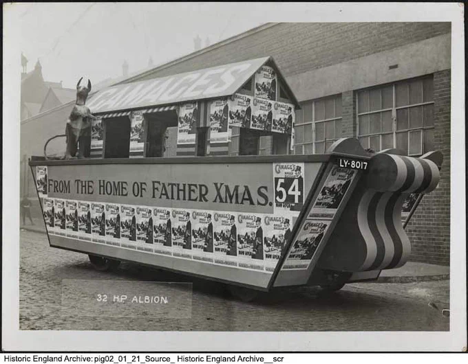 Vintage photograph showing an ark-like vehicle displaying posters and the message: 'From the Home of Father Xmas'. On the 'deck' is a hut-like structure with a pitched roof. Painted on the roof is the name: 'Gamages'.
