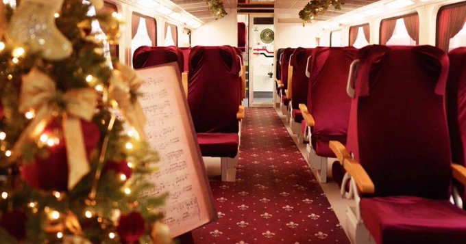 Interior of a train decorated for the holidays, featuring a Christmas tree adorned with ornaments on the left and rows of plush red seats with wooden armrests.