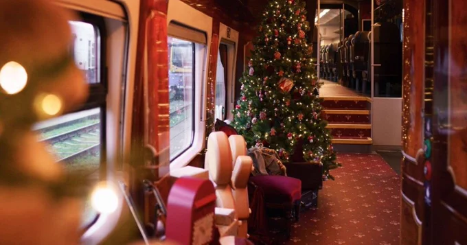 Interior of a decorated train carriage featuring a large Christmas tree and festive lights, with plush seating and gifts placed around.