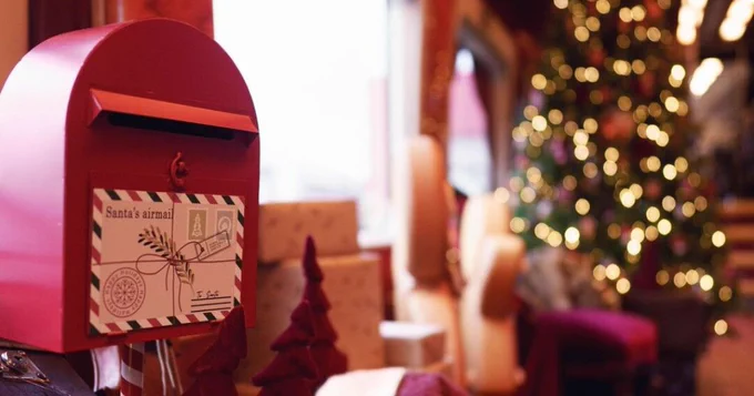 A festive scene featuring a red mailbox labeled "Santa's mail" with a decorative Christmas stamp, surrounded by gifts and a blurred Christmas tree in the background, illuminated with fairy lights.