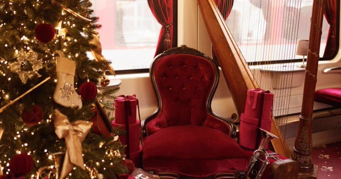 Interior of the Venice Simplon-Orient-Express train decorated for Christmas, featuring a festively adorned Christmas tree and a luxurious red velvet chair surrounded by presents.