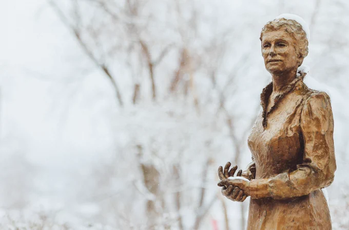 Bronze statue of Sister of Mercy Catherine McAuley standing outdoors in winter, lightly covered in snow, with bare trees in the background on the Mount Mercy campus.
