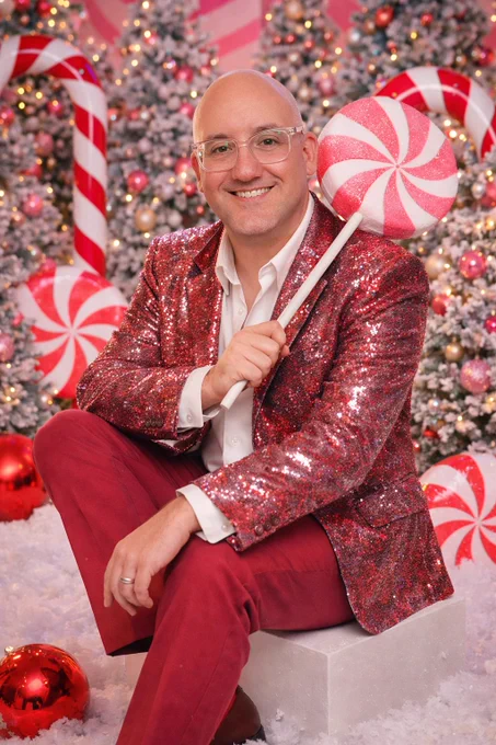 A smiling man wearing glasses and a red sequin jacket sits on a white cube in a festive Christmas studio. He holds a large pink-and-white peppermint lollipop over his shoulder, surrounded by snowy ground, decorated Christmas trees with lights, oversized candy canes, and shiny red ornaments in a bright, playful holiday setting.