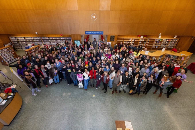 A large group of Oakland Public Library staff gathered inside a the Main library, smiling and posing for a photo. The library features bookshelves in the background and a warm, wooden interior.