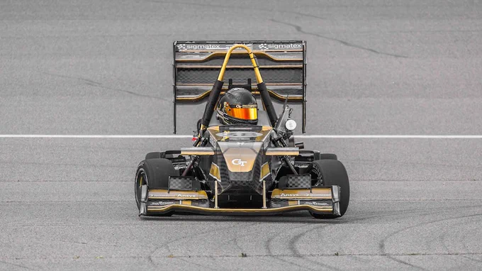 A race car with prominent Sigma Mark IV branding in black and gold colors, driven by a person wearing an orange helmet, is captured on a racing track.