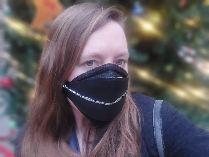 A selfie behind a Christmas tree with lights fuzzy. I'm wearing a black can99 ffp3 mask with a silver mask necklace as a mask chain. 