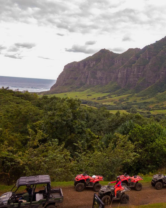 View of ATVs parked on a grassy overlook with the lush, green mountains of O'ahu in the background and a glimpse of the ocean in the distance.