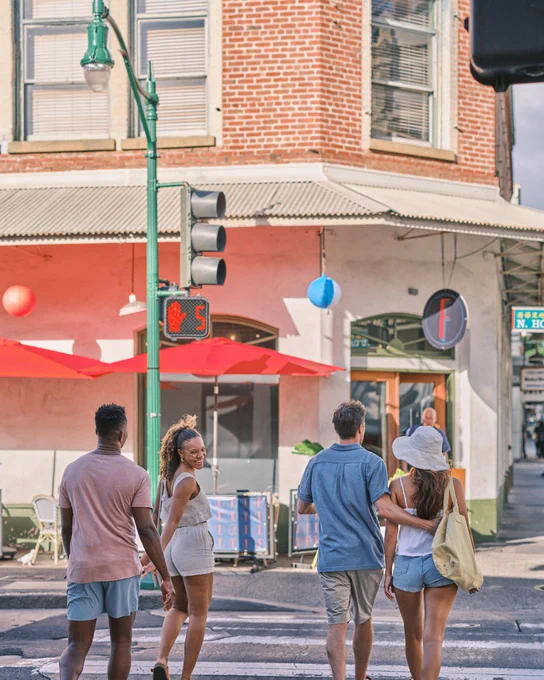 Four people crossing a street in a sunny urban setting, with traffic lights displaying the walk signal. There are colorful building facades and street lamps visible.