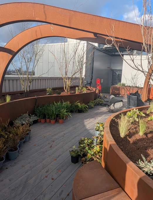 Curved rust-colored metal arches frame a rooftop garden with potted plants and wooden decking under a partly cloudy sky.