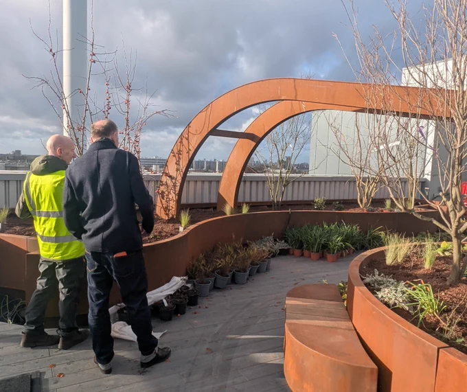 Two men, one in a yellow safety vest, inspect a rooftop garden with curved rust-colored metal planters under a partly cloudy sky.