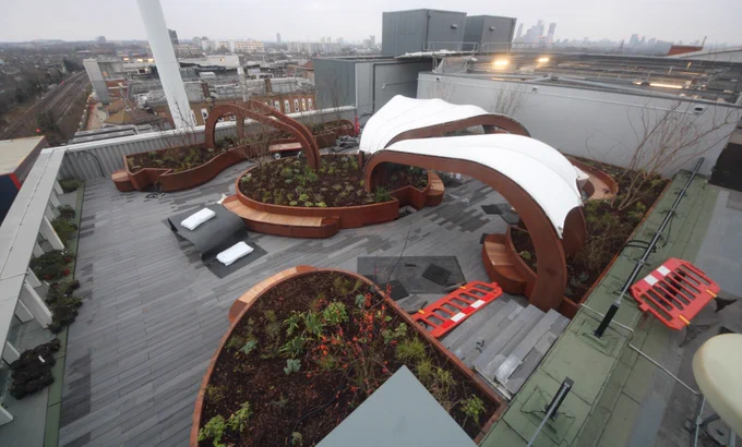 Modern rooftop garden with curved wooden benches and white fabric canopies overlooking a city skyline on a cloudy day.
