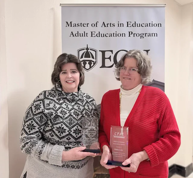 Two individuals holding awards stand in front of a banner that reads "Master of Arts in Education Adult Education Program" at ECU.