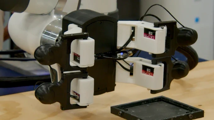 A close-up view of a robotic arm with attached grippers, featuring visible "Robotiq" labels, positioned near a worktable.