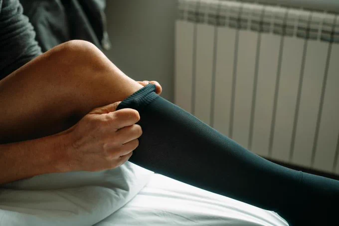 Person pulling on a compression sock while sitting on a bed.