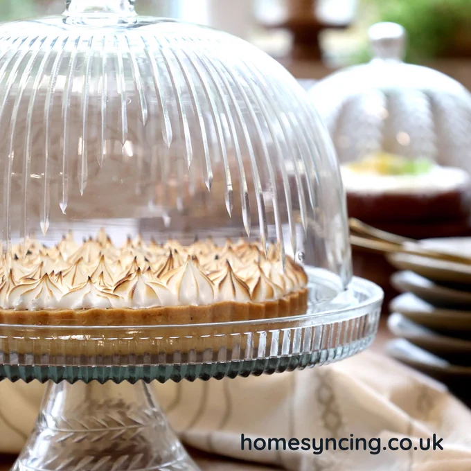 Close-up of a dessert displayed on a clear glass cake stand with a ribbed glass dome lid, showing a tart topped with toasted meringue, set on a styled table with plates and cutlery in the background, and “homesyncing.co.uk” text visible.
