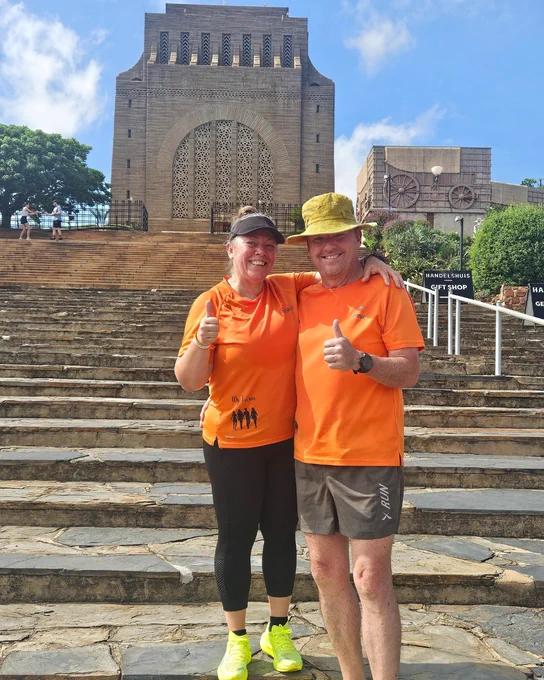 Two parkrunners in matching orange shirts smiling and giving a thumbs-up in front of the Voortrekker Monument.