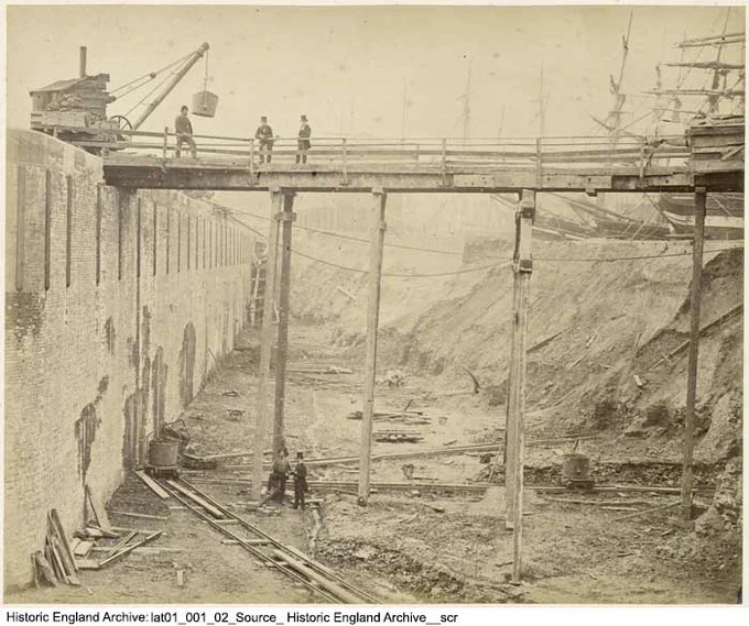 Vintage photograph of the construction of a huge trench to the side of a huge brick wall. A timber bridge spans from the top of the wall to an earth embankment. Three men stand on the bridge and two men pose in the trench. A steam crane stands next to the bridge. Several sailing ships are moored above the trench, adjacent to the embankment.