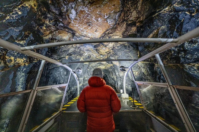 A person in a red coat boats through Standedge tunnel illuminated by lights. The exposed rock in the tunnel is coloured black and brown with seams of yellow.  