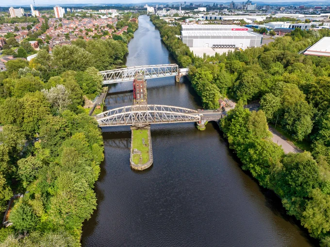 Barton Swing Aqueduct crosses a vast expanse of water surrounded by green trees. The bridge is a white metal structure. In the distance stand factories and modern buildings. 