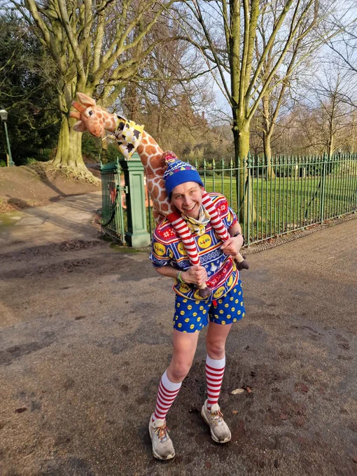 A parkrunner wearing a colorful clown costume holding a plush giraffe toy, standing in at a parkrun event. 