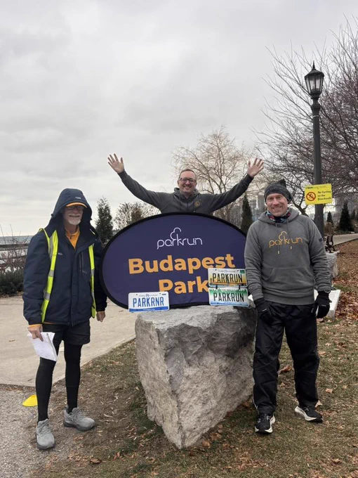 Three parkrunners, with the one at the middle with his arms up, standing beside the purple parkrun panel mounted on a rock at the Budapest Park parkrun event.