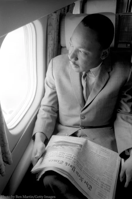 Martin Luther King Jr. seated by an airplane window, reading a newspaper in December 1963. 