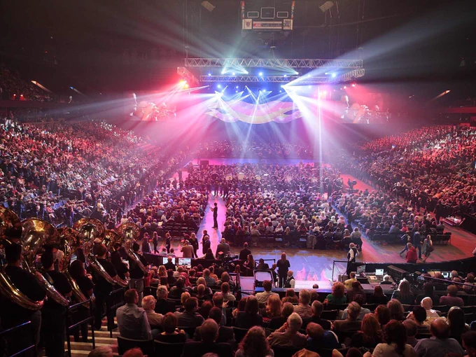 A bustling indoor concert at Van Andel Arena filled with a large audience. Bright stage lights illuminate a central performance area where musicians with various instruments prepare for the show.